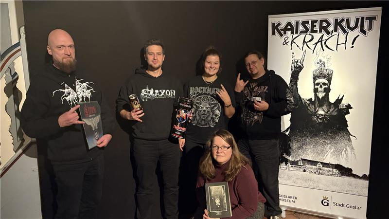 Fünf Personen vor einem Poster mit der Aufschrift 'KAISER KULT & KRACH' und einer Skelettfigur mit Krone, vier stehen, eine kniet, alle tragen dunkle Kleidung mit Bandmotiven und halten CDs oder Getränke.