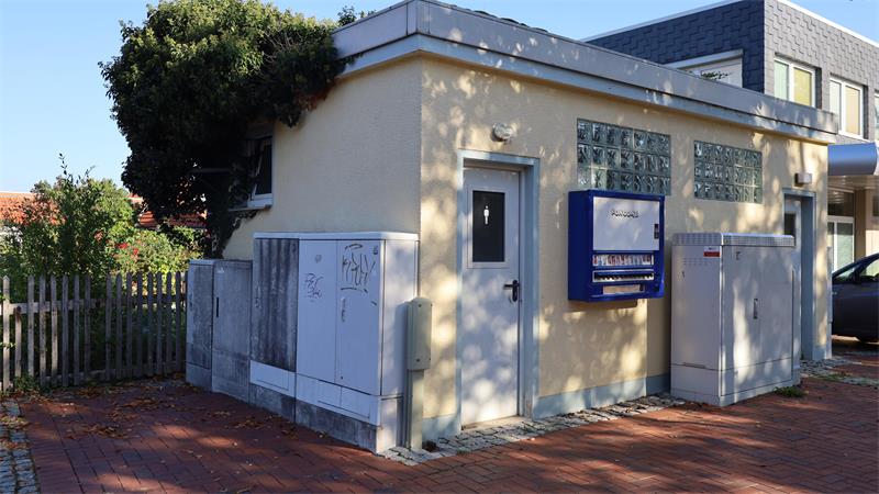 Kleines Gebäude mit weißer Tür, blauer Briefkasten und mehreren weißen Schaltkästen davor