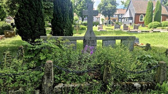 Kriegsgräberstätte mit Kreuz auf einem von Büschen und Gras überwucherten Grab auf einem Friedhof mit weiteren Gräbern und Häusern im Hintergrund