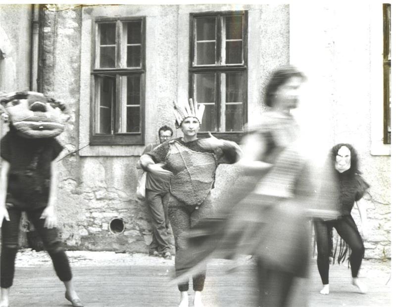 Eine Schwarz-weiß-Aufnahme zeigt verkleidete Gestalten, die sich auf einer Straße vor einem Steinhaus so stark bewegen, dass sie Bewegungsunschärfe aufweisen.
