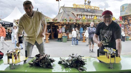 Zwei Männer spielen an einem Stand auf einem Jahrmarkt, im Hintergrund ein Gebäude mit der Aufschrift 'Bauernkate'