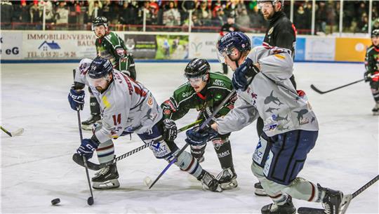 Drei Eishockeyspieler, zwei in grauen Trikots, einer im grünen Trikot, jagen den Puck.