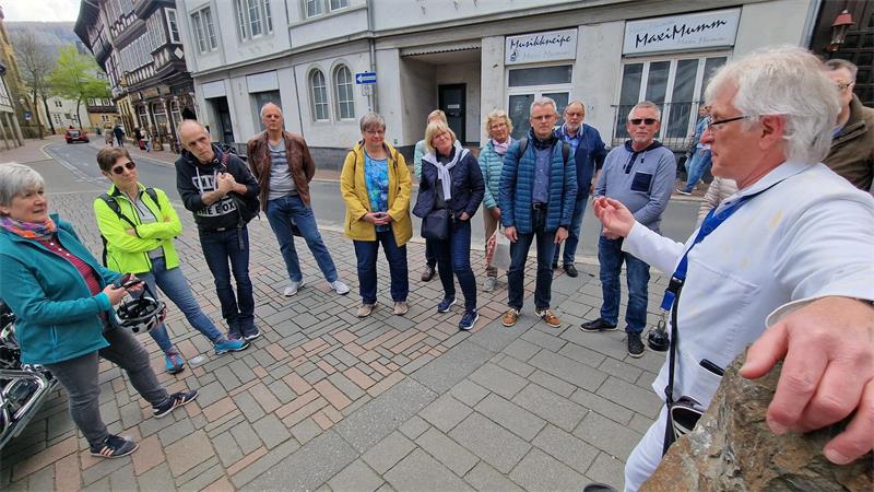 Gruppe von Menschen steht auf gepflastertem Platz und hört einem älteren Mann mit weißem Hemd zu, der sich an einen Stein lehnt.