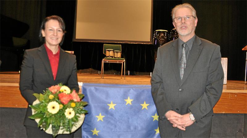 Zwei Personen in formeller Kleidung stehen vor einer Bühne mit einem Tisch, auf dem eine Europaflagge liegt, die linke Person hält einen Blumenstrauß