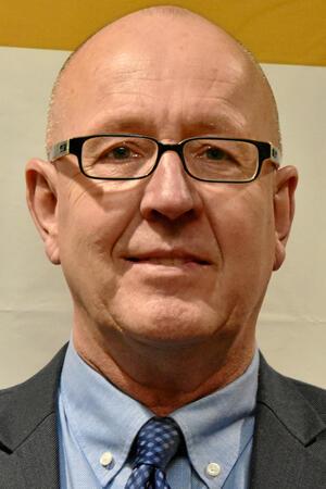 Ein Mann mit Glatze trägt einen dunkelgrauen Anzug, ein hellblaues Hemd und eine dunkelblaue Krawatte mit Muster.