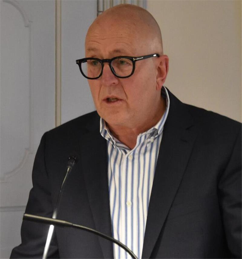 Mann mit Brille trägt gestreiftes Hemd und dunklen Anzug vor Mikrofon in Innenraum