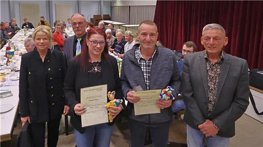 Fünf Personen stehen in einem Saal vor einem Publikum, zwei halten Urkunden und kleine Geschenke, im Hintergrund sind lange Tische mit Gästen und eine Bühne mit rotem Vorhang zu sehen.