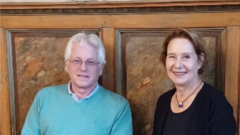 Zwei Personen sitzen vor einer holzvertäfelten Wand, eine trägt einen hellblauen Pullover, die andere ein schwarzes Oberteil mit einer Halskette