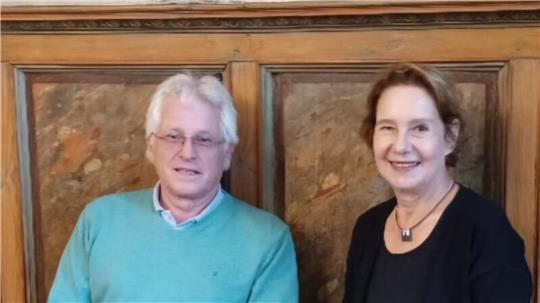 Zwei Personen sitzen vor einer holzvertäfelten Wand, eine trägt einen hellblauen Pullover, die andere ein schwarzes Oberteil mit einer Halskette