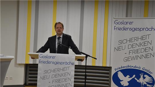 Hauke Ritz steht an einem Rednerpult mit Mikrofon vor einer Wand mit gelben senkrechten Streifen und zwei Bannern mit dem Text 'Goslarer Friedensgespräche "Sicherheit neu denken - Frieden gewinnen" und einem Logo mit zwei Tauben.