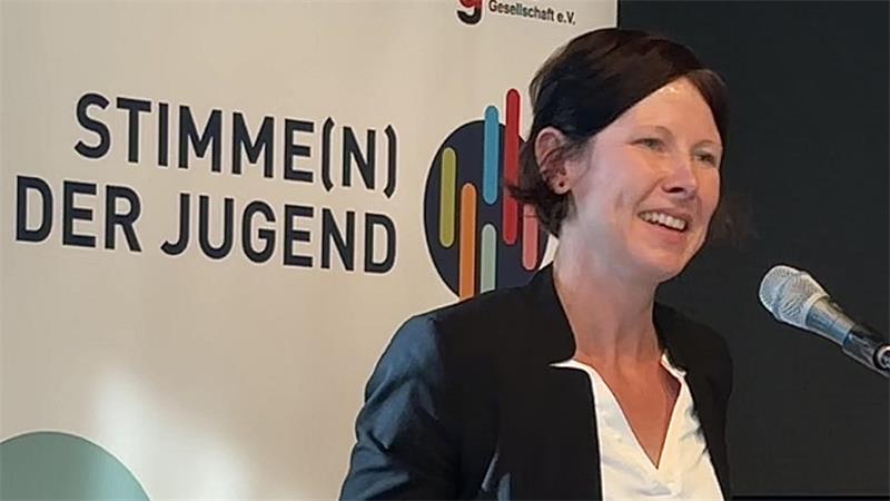 Das Foto zeigt Dr. Heike Tuchscheerer, Referentin für Politik und Geschichte bei der Deutschen Gesellschaft e.V. Links hinter ihr zeigt eine Plakatwand die Aufschrift "Stimmen(n) der Jugend".