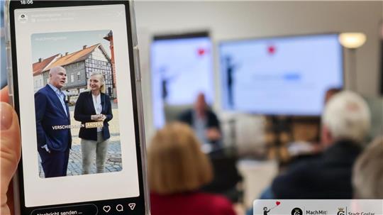 Zwei unscharfe Personen in Anzügen auf einem Smartphonebild vor einem Fachwerkhaus, im Hintergrund unscharfe Personen und ein Bildschirm in einem Raum.