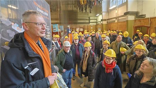 Ein Mann mit Schal und Helm in der Hand spricht zu einer großen Gruppe an Menschen mit gelben Helmen.