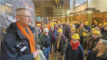 Ein Mann mit Schal und Helm in der Hand spricht zu einer großen Gruppe an Menschen mit gelben Helmen.