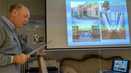 Mann hält Papier und steht vor einer Leinwand mit einer Präsentation über Architektur in einem Raum mit Sofa und Stuhl