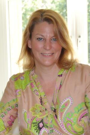 Frau mit schulterlangem, gewelltem Haar trägt ein hellbraunes Hemd mit grünem und pinkem Paisley-Muster und steht vor einem Fenster