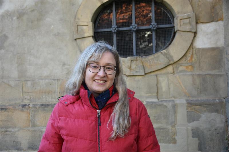 Eine Frau in roter Jacke steht vor einer Sandsteinmauer mit vergittertem Fenster. 