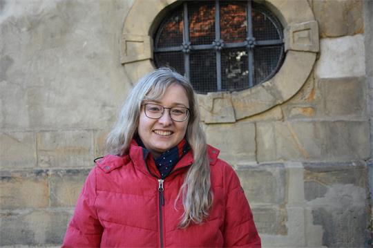 Eine Frau in roter Jacke steht vor einer Sandsteinmauer mit vergittertem Fenster. 