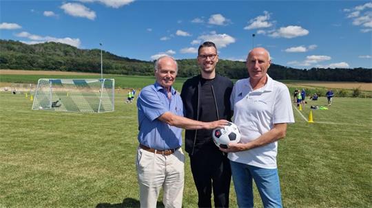 Drei Männer stehen auf einem Fußballfeld, zwei von ihnen halten gemeinsam einen Fußball, im Hintergrund sind weitere Spieler und ein Tor zu sehen.