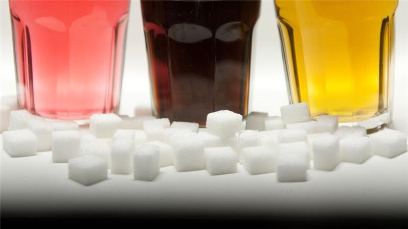 Softdrinks erhöhen Risiko für Diabetes und Herzkrankheiten Drei Gläser gefüllt mit roter Limonade, Cola und einem Energy-Drink neben Zuckerwürfeln auf einem Tisch