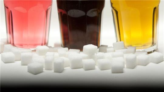 Drei Gläser gefüllt mit roter Limonade, Cola und einem Energy-Drink neben Zuckerwürfeln auf einem Tisch