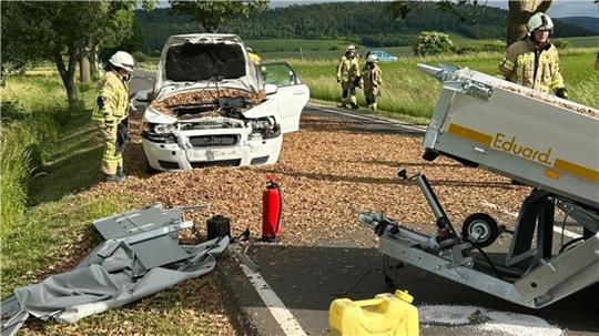 Unfallstelle auf Landstraße mit beschädigtem weißem Auto, umgestürztem Anhänger mit Aufschrift 'Eduard', mehreren Feuerwehrleuten und verstreutem Ladegut