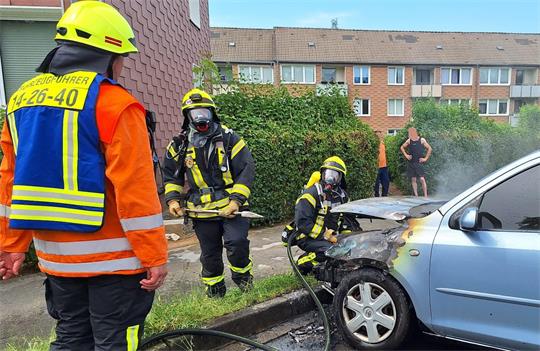 Das Foto zeigt drei Feuerwehrwehrleute mit Löschmitteln vor einem defekten Auto.