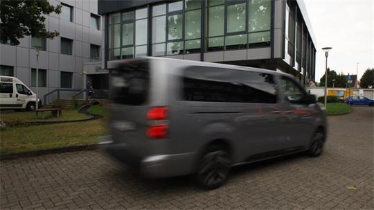 Silberner Van fährt auf gepflastertem Hof vor einem modernen Bürogebäude mit großen Fenstern