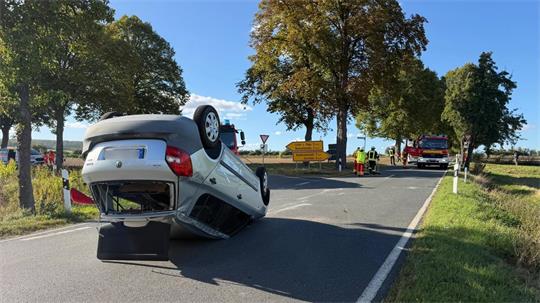 Ein Auto liegt auf einer Landstraße auf dem Dach. Feuerwehrautos und -leute stehen im Hintergrund.