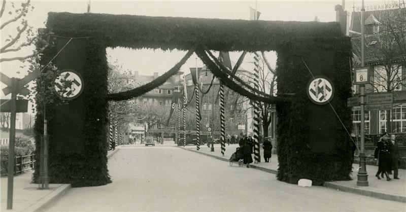 Straße mit einem großen Torbogen, der mit Tannenzweigen geschmückt ist und an den Seiten Hakenkreuzsymbole zeigt.