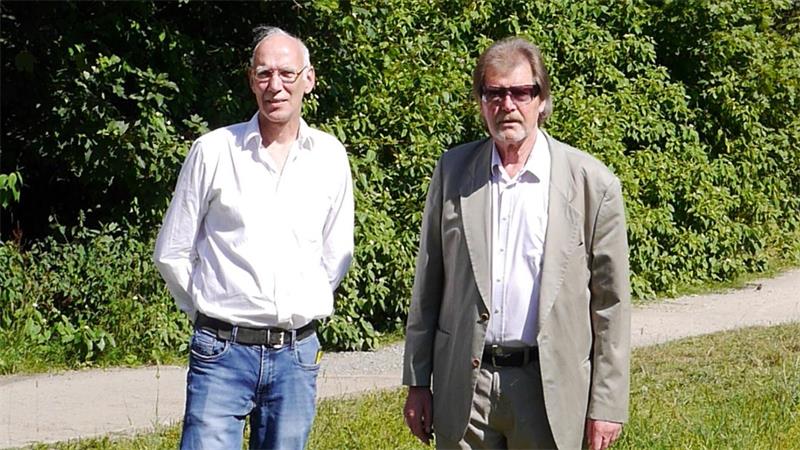 Zwei Männer stehen auf einem Grasweg vor dichtem grünem Gebüsch, der linke trägt ein weißes Hemd und Jeans, der rechte einen hellen Anzug mit weißem Hemd und Sonnenbrille