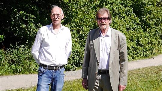 Zwei Männer stehen auf einem Grasweg vor dichtem grünem Gebüsch, der linke trägt ein weißes Hemd und Jeans, der rechte einen hellen Anzug mit weißem Hemd und Sonnenbrille