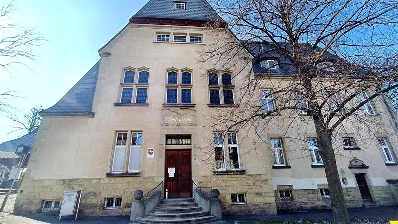 Fassade eines historischen Gebäudes mit steilem Schieferdach, mehreren Fenstern und einem Eingang mit Treppe, davor ein kahler Baum