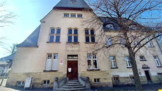 Fassade eines historischen Gebäudes mit steilem Schieferdach, mehreren Fenstern und einem Eingang mit Treppe, davor ein kahler Baum