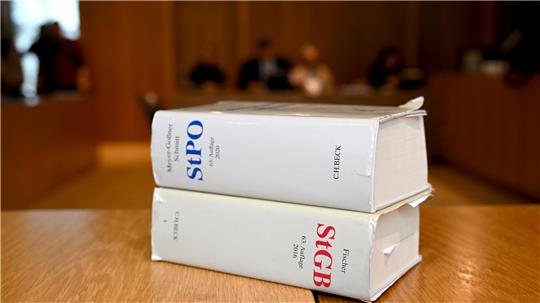 Zwei dicke Gesetzesbücher mit den Titeln 'StPO' und 'StGB' liegen auf einem Holztisch in einem Gerichtssaal