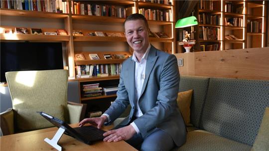 Ein jüngerer Mann im blau-grauen Anzug sitzt lächelnd in einer Bibliothek auf einem Sofa und hält sein Laptop, das auf einem Tisch vor ihm steht.