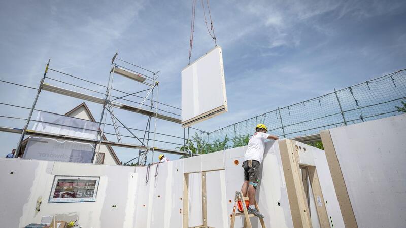 Weniger Fertighäuser gebaut Ein Bauarbeiter steht auf einer Leiter und arbeitet im Rohbau eines Fertighauses, während gerade ein weiteres Wandelement von einem Kran heruntergelassen wird.