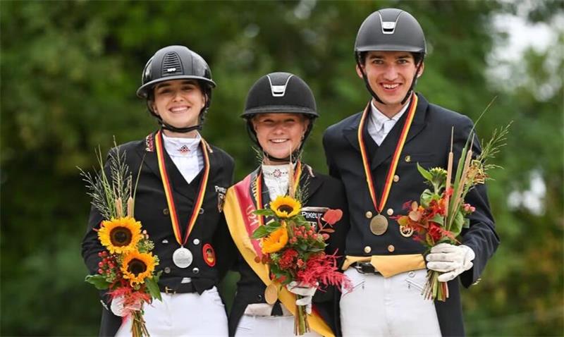 Drei junge Reiter in schwarzer Reitkleidung und weißen Reithosen mit Medaillen und Blumensträußen lächelnd vor grünem Hintergrund