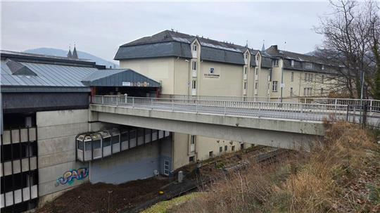 Brücke verbindet zwei Gebäudeteile über einem Abhang mit kahlen Bäumen und brauner Vegetation im Vordergrund