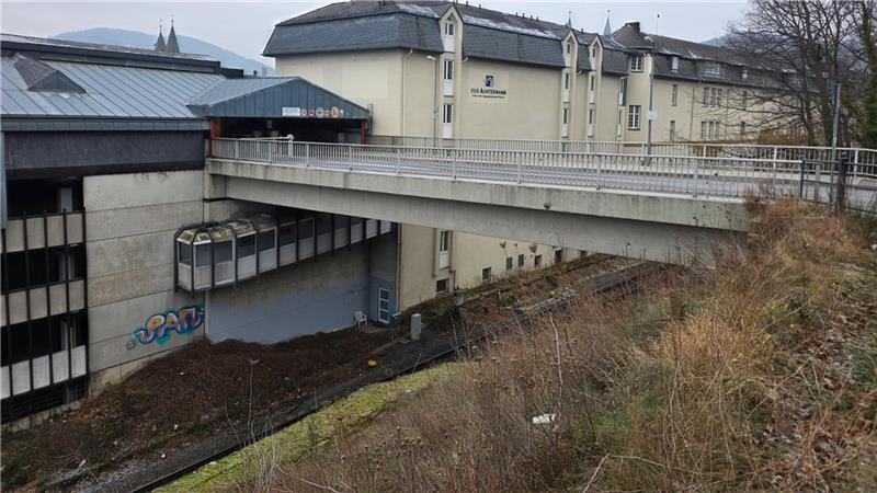 Brücke verbindet zwei Gebäudeteile über einem Abhang mit kahlen Bäumen und brauner Vegetation im Vordergrund