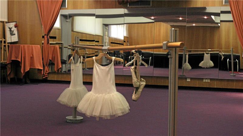 Ballettkleider und Ballettschuhe hängen an einer Stange vor einem Spiegel in einem Tanzstudio mit lila Teppichboden und orangen Vorhängen