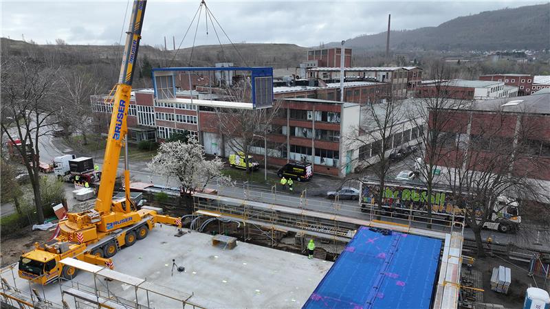 Kran im Einsatz: Spektakulärer Ausbau der Grundschule Oker Baukran hebt blauen Container über Baustelle mit Betonplatte in städtischer Umgebung mit Gebäuden und Bäumen