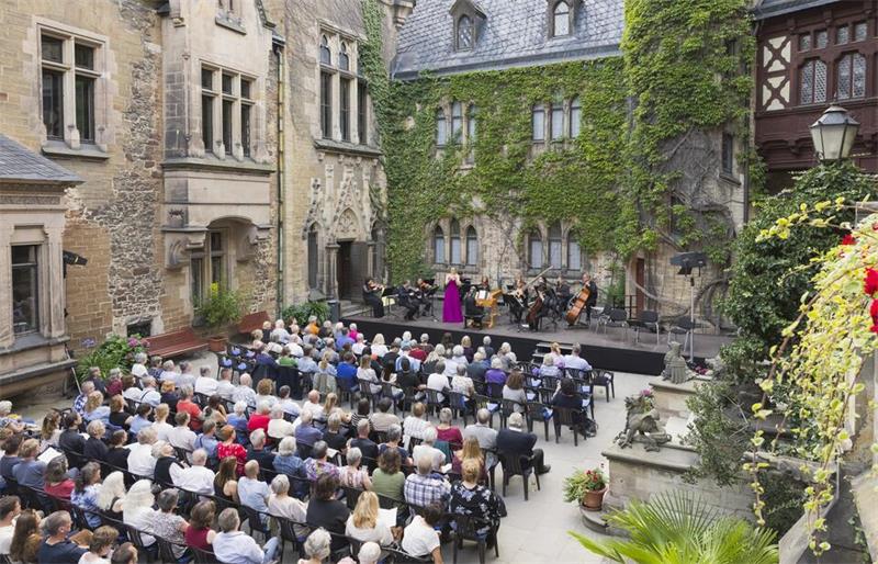 In einem herrschaftlichen Innanhof mit efeubewachsenen historischen Gebäudefassaden sitzen Menschen auf Stühlen vor einer Bühne, auf der Musiker mit klassischen Instrumenten musizieren.