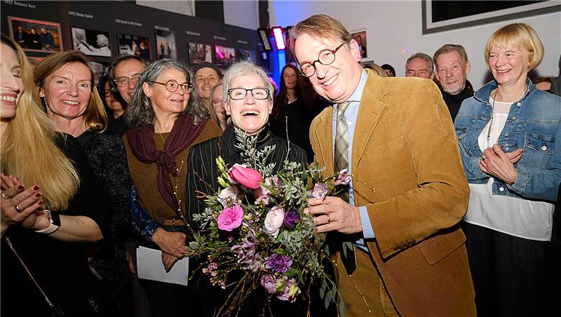 Ein Mann übergibt einer Frau einen bunten Blumenstrauß in pink, rosa und violett; die Beschenkte lacht in die Kamera, um die beiden stehen viele lächelnde Menschen herum. 