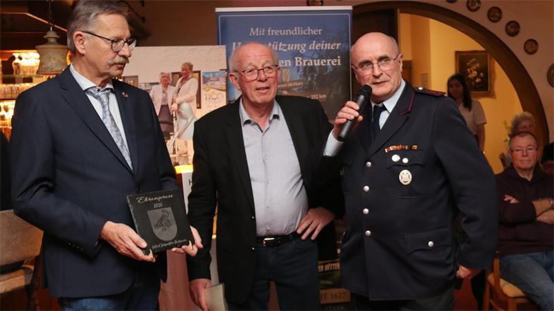 Drei Männer stehen nebeneinander, einer hält ein Mikrofon, ein anderer ein Wappen, im Hintergrund Banner und weitere Personen in einem Raum mit warmem Licht.