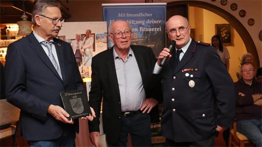Drei Männer stehen nebeneinander, einer hält ein Mikrofon, ein anderer ein Wappen, im Hintergrund Banner und weitere Personen in einem Raum mit warmem Licht.
