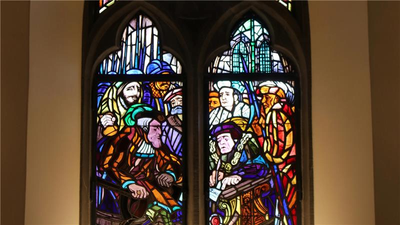 Buntes Glasfenster mit mehreren Personen in historischer Kleidung, die an einem Tisch sitzen