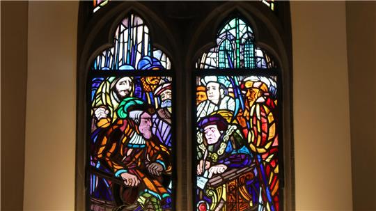 Buntes Glasfenster mit mehreren Personen in historischer Kleidung, die an einem Tisch sitzen