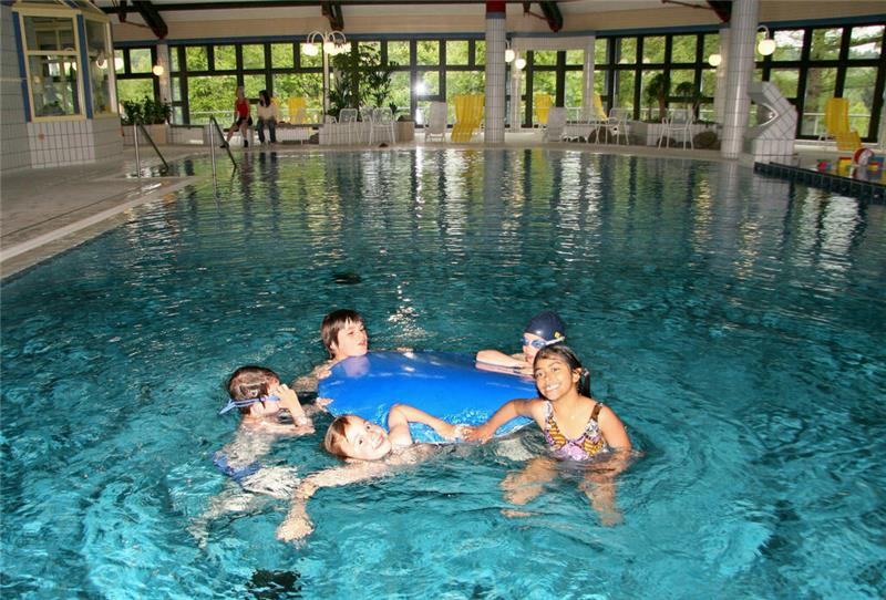 Mehrere Kinder spielen und schwimmen in einem Hallenbad mit großen Fenstern und gelben Stühlen am Beckenrand.