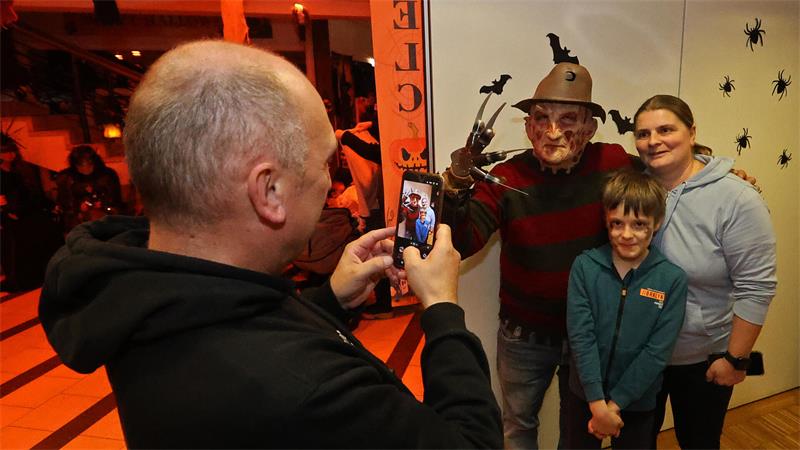 Ein Mann fotografiert eine Frau und ein Kind zusammen mit einer Person in einem Freddy-Krueger-Kostüm mit braunem Hut und Metallklauen vor einer weißen Wand mit schwarzen Spinnenaufklebern.